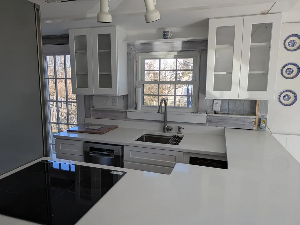 White shaker kitchen with glass-front wall cabinets, sink under a window and black glass cooktop in front.